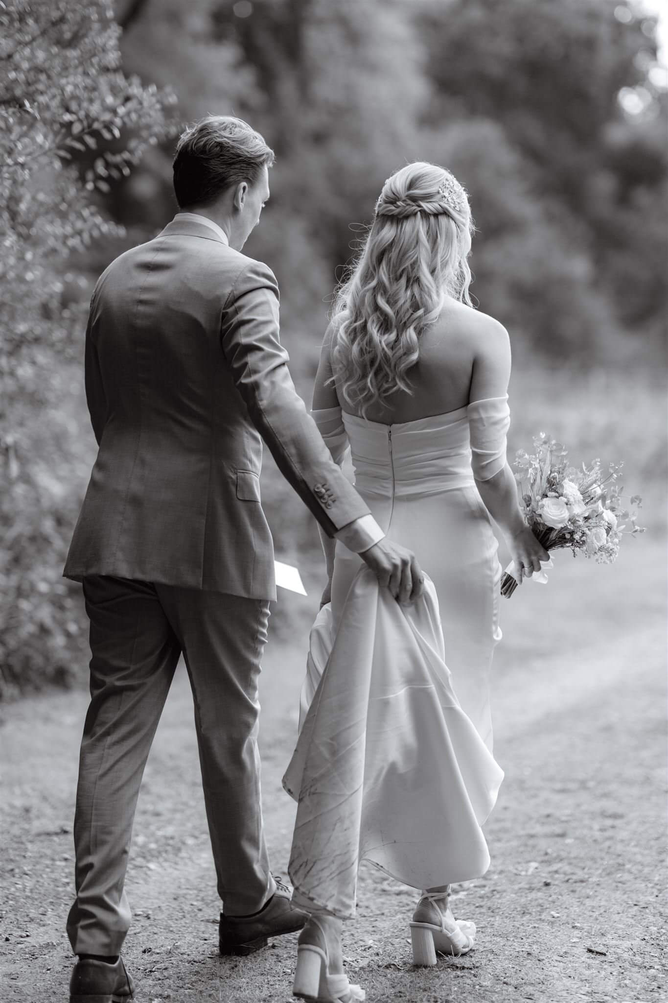 Groom holding up brides dress while walking through a field. Hair created by Surrey based wedding hairstylist, Lucy Hayward.